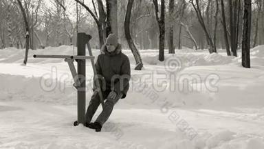 穿蓝色羽绒服带头罩的人在冬季公园前景B W模拟器上做腿部运动